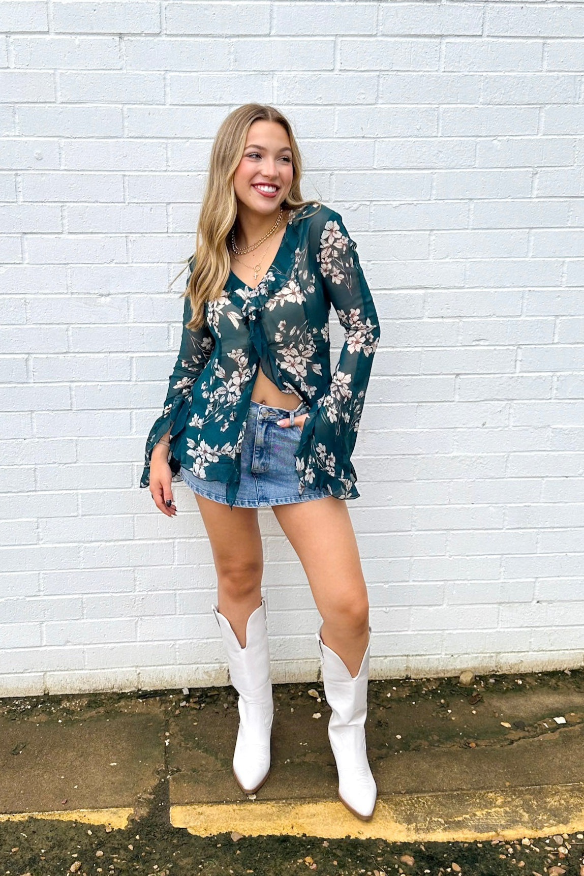 Woman wearing a floral top and denim shorts with white boots against a white brick wall.