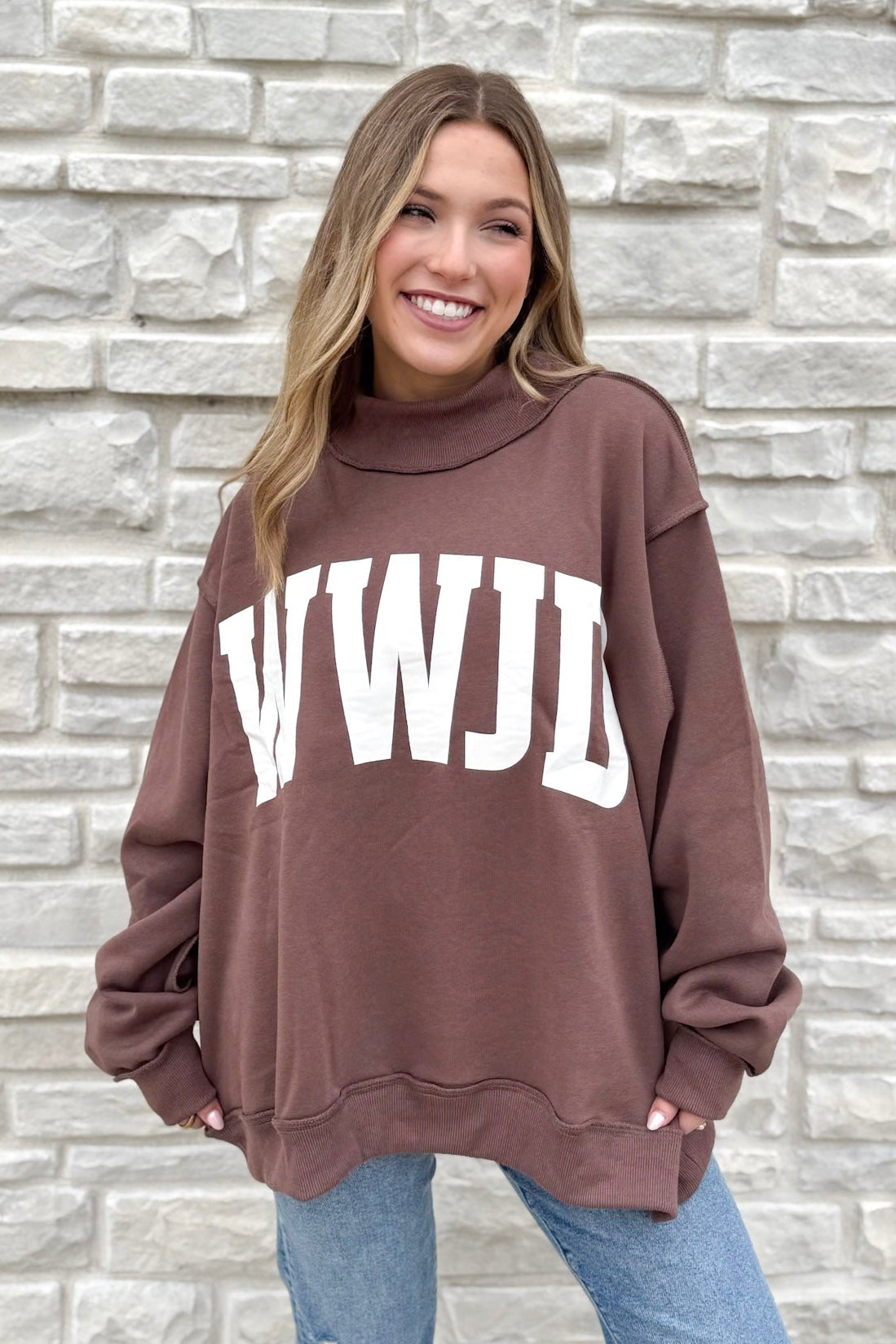 A smiling woman with long, light brown hair wears the WWJD/Amen Mockneck sweatshirt with bold white WWJD print, standing in front of a light stone wall. Her hands are in her pockets and she pairs it with blue jeans.