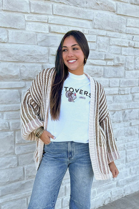 Woman wearing a cardigan, t-shirt, and jeans standing against a light stone wall.