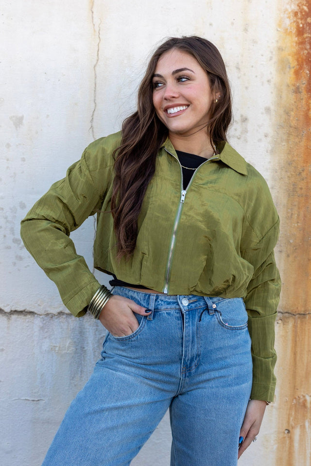 Woman wearing a green jacket and blue jeans standing against a textured wall.