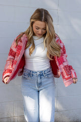 Woman wearing a red patterned jacket, white top, and blue jeans against a white wall.