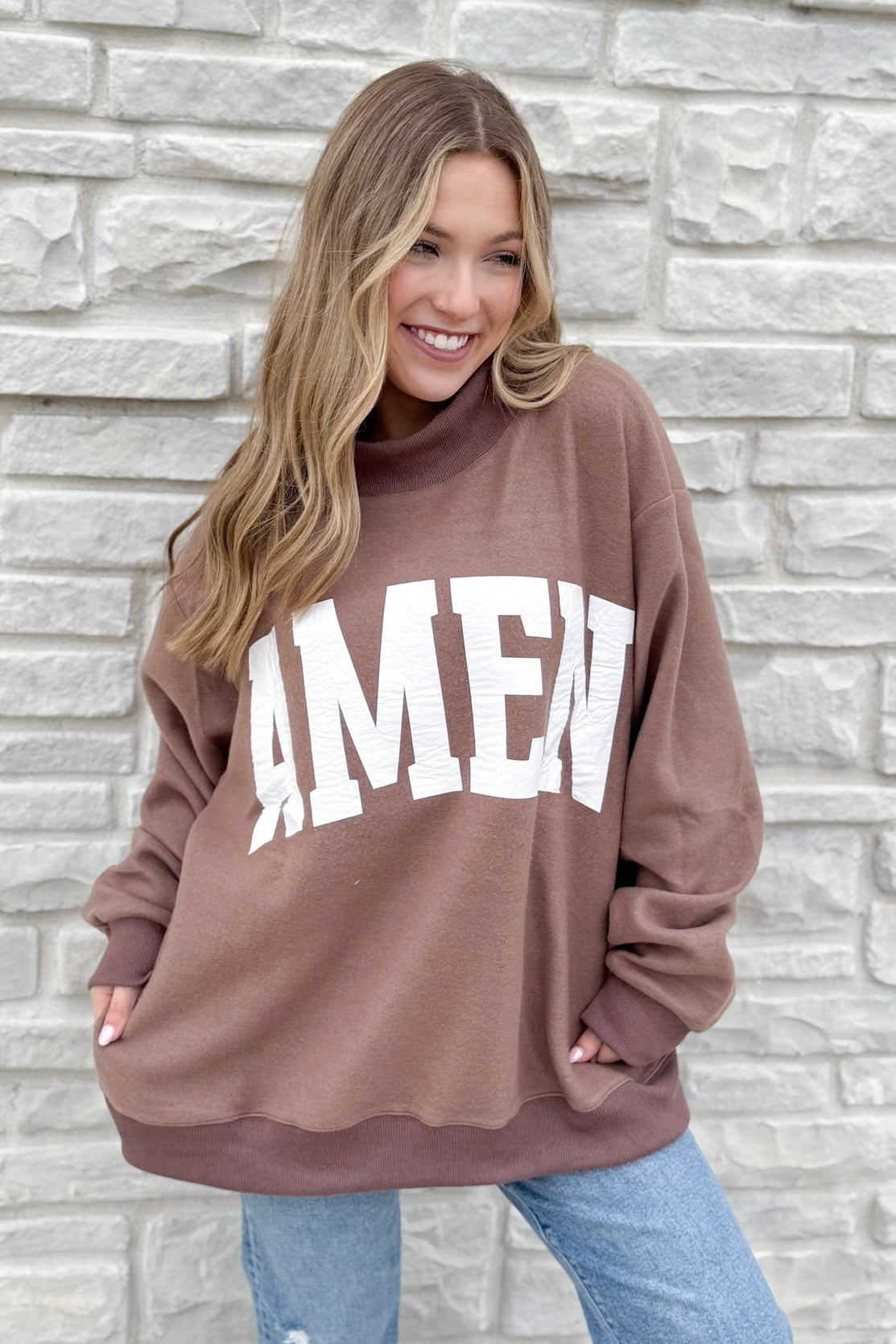 A smiling woman with long, wavy blonde hair stands by a light stone wall, wearing the WWJD/Amen Mockneck—a loose brown sweatshirt with "AMEN" in bold white letters—paired with blue jeans.