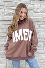 A smiling woman with long, wavy blonde hair stands by a light stone wall, wearing the WWJD/Amen Mockneck—a loose brown sweatshirt with "AMEN" in bold white letters—paired with blue jeans.