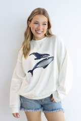 Woman wearing a white sweatshirt with a whale design against a plain background